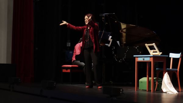 La récitante, bras tendus, devant le piano à queue et le décor du spectacle