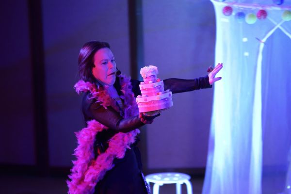 La récitante, boa rose aux épaules, présente un gâteau à trois étages sur une scène éclairée de bleu et de rose